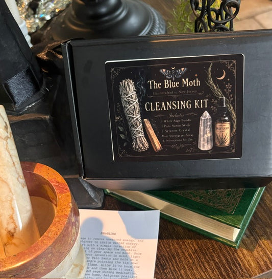 A black box labeled 'The Blue Moth Cleansing Kit' sits on a wooden surface with a candle, book, and printed card nearby. The box front shows illustrated white sage, a crystal, and a jar, and the scene appears styled as a tabletop product presentation.